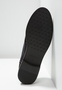 Schwarzer Schuh mit einer glatten Gummisohle, die ein strukturiertes Muster aufweist, weiches Obermaterial aus Stoff und sichtbarem Markenlogo auf der Sohle, schräger Blick.