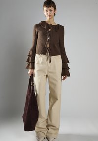 Brauner gestrickter Pullover mit einem Rüschenausschnitt, vorne gebunden; hellbeige weit geschnittene Hose; dunkelburgunderfarbene Wildledertasche; helle Schuhe.