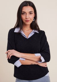 Pull noir en maille texturée avec un col en V, superposé sur une chemise à col bleu clair. Les manches sont retroussées, montrant les poignets de la chemise.