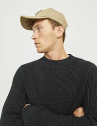 Beige színű baseball sapka elöl varrott részletekkel, egy textur ált fekete pulóverrel viselve, amely környakas és hosszú ujjú.