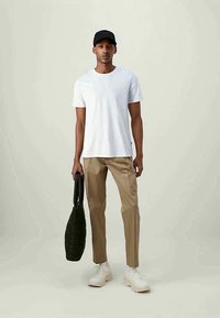 Man wearing white t-shirt, beige pants, white sneakers, black cap, holding black quilted bag against plain light background.