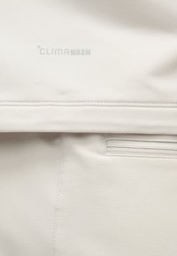 Nahaufnahme eines weißen Stoffs mit genähten Nähten und einer kleinen Reißverschlusstasche, auf der oberen Partie mit dem aufgedruckten "CLIMAWARM"-Logo.