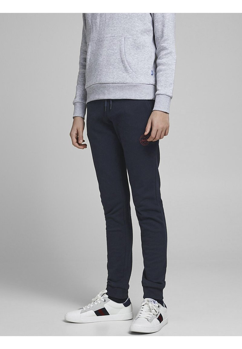 Šedá mikina s přední kapsou, námořnické tepláky s vyšitým logem a bílé tenisky s červenými a námořnickými akcenty.