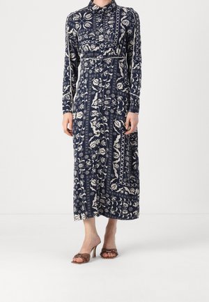 Femme portant une robe longue bleue marine et blanche à motifs floraux, boutonnée, manches longues, avec des sandales marron à talons hauts.