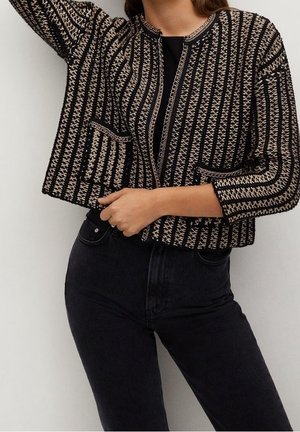 Femme portant un cardigan tricoté à rayures noires et beiges avec des poches à l’avant, par-dessus un haut noir et un pantalon noir.