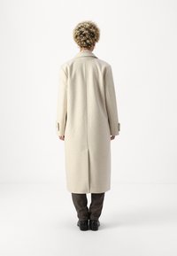 Cappotto di lana beige con una silhouette lunga e dritta. Presenta un colletto a revers e polsini con bottoni. Texture liscia e design lungo fino al ginocchio.