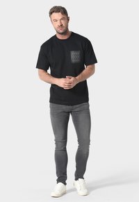 Črna bombažna majica s potiskanim žepom, v kombinaciji s sivimi ozkimi skinny jeans hlačami in belimi supergami. Model stoji pred nevtralno ozadjem.