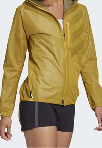 Geel lichtgewicht jack met een ritssluiting aan de zak, een mesh textuur en elastische manchetten. Zwarte korte broek met grijze strepen aan de zijkanten.