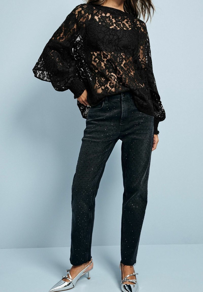 Top in pizzo nero con maniche lunghe e ampie, abbinato a jeans in denim nero con borchie argentate. Il modello indossa tacchi a punta argentati.