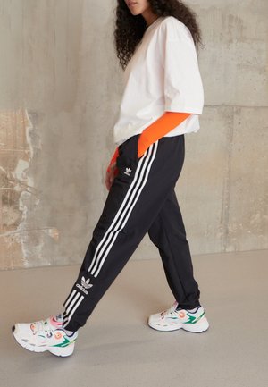 Femme marchant à l'intérieur portant un T-shirt blanc oversized, un pantalon de survêtement Adidas noir avec des bandes blanches, un sous-pull orange vif à manches longues et des baskets colorées.