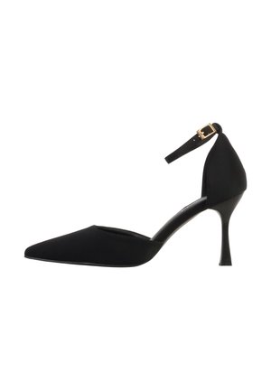 Zapato de tacón alto negro con punta puntiaguda, tacón delgado y correa en el tobillo con hebilla dorada, mostrado de lado sobre un fondo blanco.