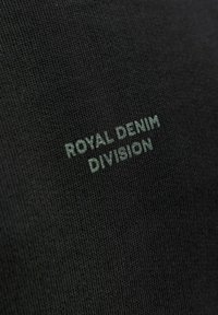 Czarna tkanina z teksturowanym splotem, na którym wytłoczone jest napis "ROYAL DENIM DIVISION" w kontrastowym, stonowanym zielonym kolorze.