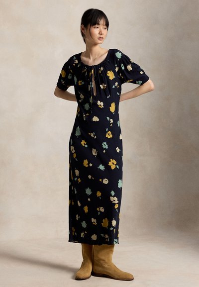 Γυναίκα με μαύρο midi φόρεμα με floral σχέδιο και κοντά φουσκωτά μανίκια, καθώς και ταμπά σουέντ μποτάκια μέχρι τον αστράγαλο, στέκεται μπροστά σε μια απλή μπεζ φόντο.