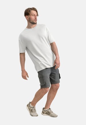 Witte, kortaf t-shirt gecombineerd met grijze cargoshorts. Schoenen zijn lichtgrijs met witte accenten. Model staat in een lichte hoek.