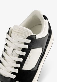Black and white sneaker featuring a leather and textile upper, rounded toe, thick white laces, and textured accents on the side.