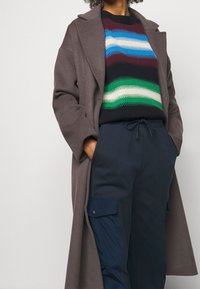 Brun ullkappa över en svart och flerfärgad randig tröja, tillsammans med marinblå sweatpants med cargo-fickor och dragsko.