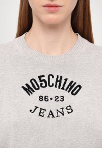 Suéter cinza claro com um decote canelado. Texto bordado em preto com a frase "MOSCHINO JEANS 86-23" em destaque na frente.