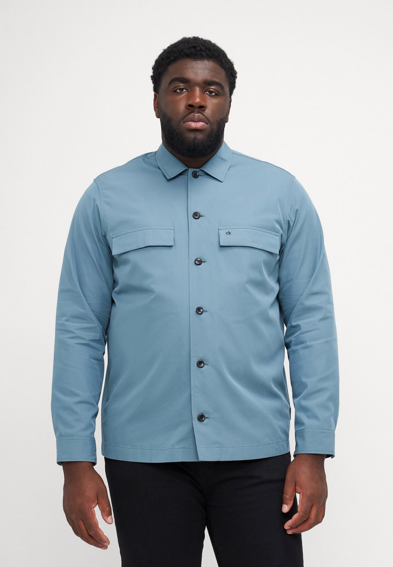 Camicia azzurro chiaro a maniche lunghe con chiusura a bottoni, due tasche sul petto e una texture liscia. Presenta un colletto e bottoni neri.