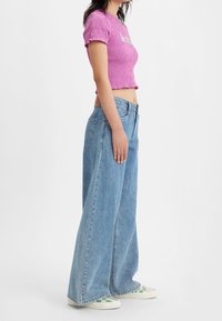 Kvinna i en lila kort topp, högmidjade vida blå jeans och vita sneakers med grön rutmönster, stående i profil.