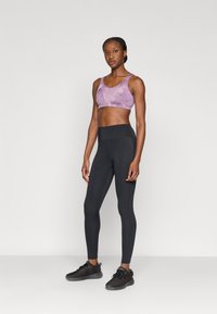 Ανοιχτό μωβ αθλητικό σουτιέν με λείο φινίρισμα, συνδυασμένο με μαύρο κολάν υψηλής μέσης και μαύρα αθλητικά παπούτσια, σε ουδέτερο φόντο.