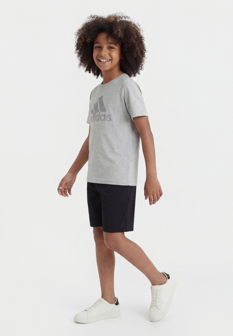 Χαμογελαστό παιδί με σγουρά μαλλιά περιπατεί, φορώντας γκρι μπλουζάκι Adidas, μαύρο σορτς και λευκά αθλητικά παπούτσια σε απλό φόντο.