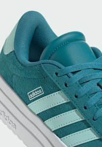 Turkos mockasinsneaker med läderdetaljer och tre vita ränder. Har en texturerad tung och snörning, robust vit sula.