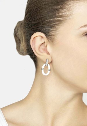White ceramic earrings with a U-shape design, featuring silver accents and small clear crystals. Displayed on a model's ear.