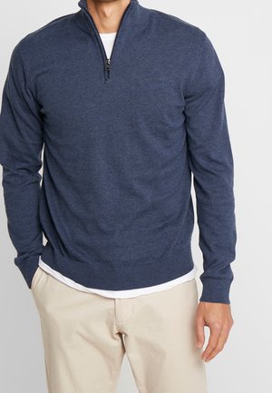 Mann trägt marineblauen Pullover mit Viertelreißverschluss über weißem Hemd und beigen Hosen, steht mit einer Hand in der Tasche vor einfarbigem Hintergrund.