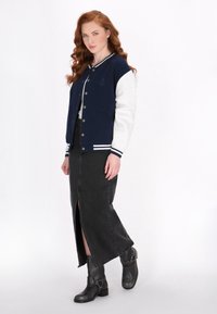 Jachetă varsity bleumarin și gri cu dungi albe, asortată cu o fustă din denim neagră cu slit frontal și bocanci negri cu accente.