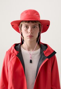 Chapeau seau rouge avec un large rebord et un cordon ajustable rose. Porté avec une veste rouge dotée d'une doublure grise et un t-shirt de couleur claire en dessous.