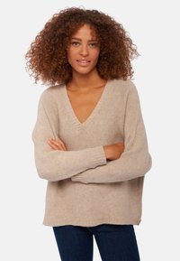 Beige gestrickter Pullover mit V-Ausschnitt, langen Ärmeln und gerippten Bündchen. Weiche Textur und lässige Passform. Über dunkelblauen Jeans getragen.