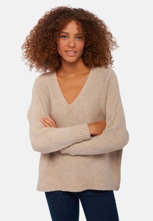 Beige gestrickter Pullover mit V-Ausschnitt, langen Ärmeln und gerippten Bündchen. Weiche Textur und lässige Passform. Über dunkelblauen Jeans getragen.