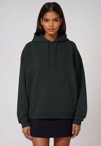 Sudadera verde oscuro hecha de tejido suave, con un bolsillo delantero, capucha con cordón y puños y dobladillo acanalados. Usada sobre una falda oscura.