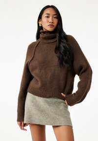 Kvinde iført en brun turtleneck sweater og en lys grå mini nederdel, poserer med den ene hånd på hoften mod en ensfarvet hvid baggrund.