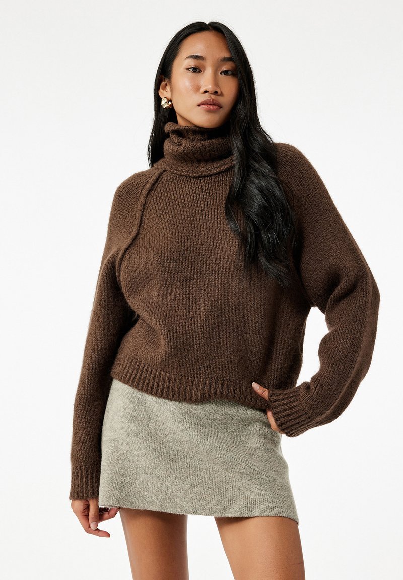 Kvinde iført en brun turtleneck sweater og en lys grå mini nederdel, poserer med den ene hånd på hoften mod en ensfarvet hvid baggrund.