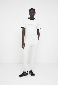 Λευκό T-shirt με μαύρο περιθώριο και το κείμενο "ORIGINALS", σε συνδυασμό με ανοιχτό γκρι υφασμάτινη φόρμα και μαύρα αθλητικά παπούτσια με λευκές λεπτομέρειες.