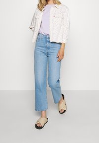 Veste-chemise blanche à motif floral, t-shirt lilas, jean large bleu clair et sandales beige à brides croisées avec semelles noires.