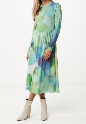 Kobieta ubrana w sukienkę z długim rękawem do połowy łydki, w stylu tie-dye w odcieniach zieleni, niebieskiego i bieli, zestawioną z białymi botkami za kostkę.