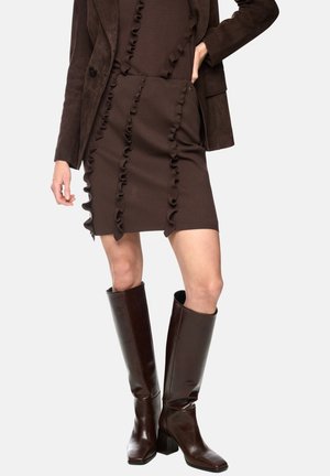 Woman wearing a dark brown ruffled skirt and top, paired with a matching brown blazer and knee-high dark brown leather boots.