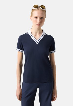 Kobieta ubrana w granatową koszulkę polo z krótkim rękawem, z białym pasiastym kołnierzykiem w kształcie litery V i mankietami, granatowe spodnie oraz białe okulary przeciwsłoneczne na głowie.