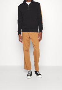Felpa nera con zip e colletto alto, maniche lunghe e strisce laterali arancioni, abbinata a pantaloni beige a gamba dritta e sneakers nere.