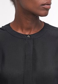 Černé tričko s kulatým výstřihem, zapínáním na jeden knoflík a hladkou texturou látky. Jednoduchý, minimalistický design bez vzorů.