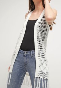 Offene, gestrickte weiße Weste mit einem langen, fließenden Design. Mit Fransenverzierungen am Saum und einem dekorativen Rautenmuster an den Seiten.