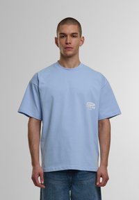 Tricou oversize de culoare albastru deschis, din bumbac, cu guler rotund. Prezintă un mic logo imprimat alb pe pieptul stâng. Mâneci scurte și largi.