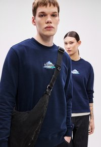 Tengerészkék pamut pulóver hímzett "Tommy Jeans" logóval, környakas, bordázott mandzsettákkal, fekete szövet oldaltáskával viselve.