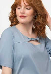 Vaaleansininen lyhythihainen paita, jossa on pyöreä pääntie ja ainutlaatuinen etuleikkaus. Valmistettu pehmeästä materiaalista, jossa on sileä pinta.