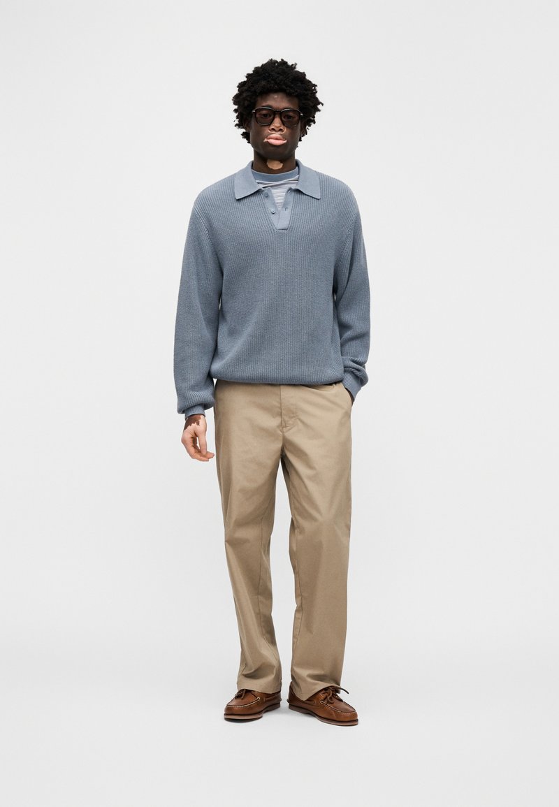 Lyseblå pullover med struktureret strik og krave, kombineret med beige bukser og brune loafers. Modellen bærer sorte, rektangulære briller.