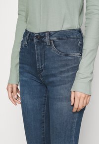 Jeans de mezclilla azul oscuro con un diseño ajustado, estilo de cinco bolsillos y cierre de botón. Se llevan con una blusa de manga larga ribeteada de color verde claro.
