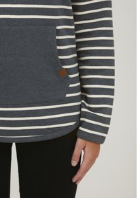 Camiseta de manga larga a rayas en tonos navy y blanco, hecha de una tela suave, con un bolsillo en el frente y un pequeño parche de logo marrón. Con dobladillo redondeado.