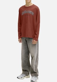 T-shirt rouge à manches longues avec "JACK & JONES" imprimé en noir sur la poitrine, associé à un jean gris ample et des chaussures de sport blanches.